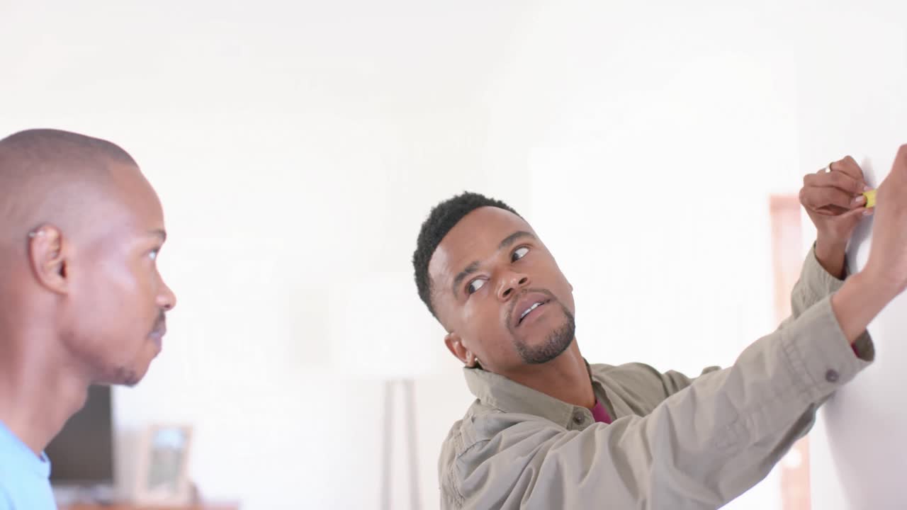 Happy african american gay male couple using tape measure and making mark on wall, slow motion