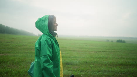 vista lateral de una mujer con un impermeable verde que lleva una bolsa de color menta y un paraguas, caminando por un campo cubierto de hierba bajo un cielo brumoso, mirando hacia la distancia, rodeada de vegetación abierta y cielo nublado