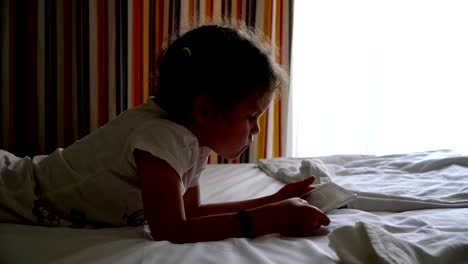 little girl plays on the bed in the tablet.