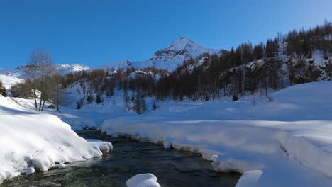 Drones-Volando-A-Baja-Altitud-Sobre-Un-Arroyo-De-Montaña-Que-Fluye-A-Través-De-Un-Paisaje-Nevado-En-Un-Día-Soleado,-Alpes-Italianos