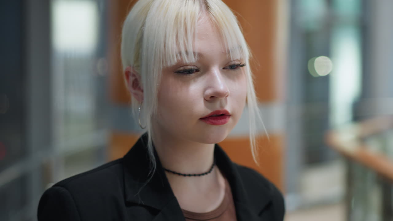 Blonde model posing stylishly indoors near glass window with hand on head and elegant posture as city lights reflect outside creating modern urban evening atmosphere inside shopping mall