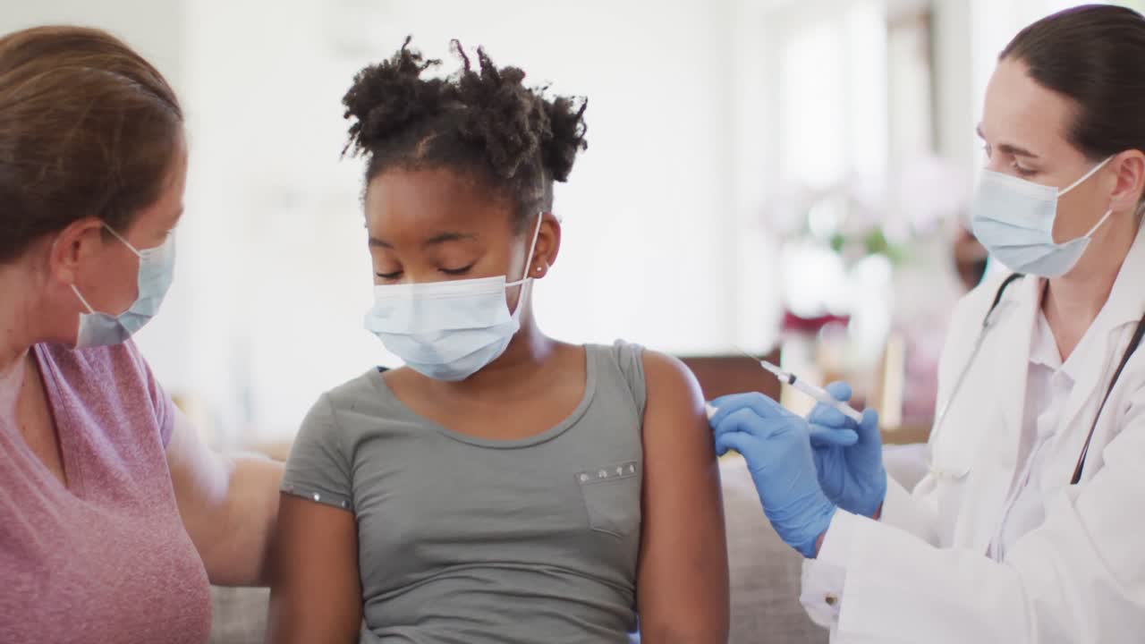 African american girl with caucasian mother and female doctor wearing face masks, vaccinating