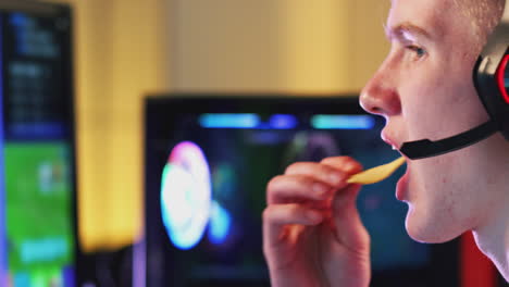 niño adolescente con auriculares comiendo papas fritas mientras juega en casa usando pantallas de computadora duales