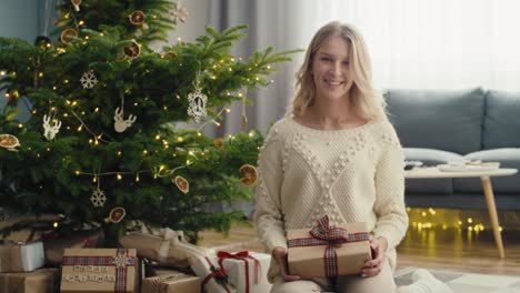 Portrait-of-caucasian-woman-sitting-next-to-Christmas-tree-and-holding-a-gift-box.