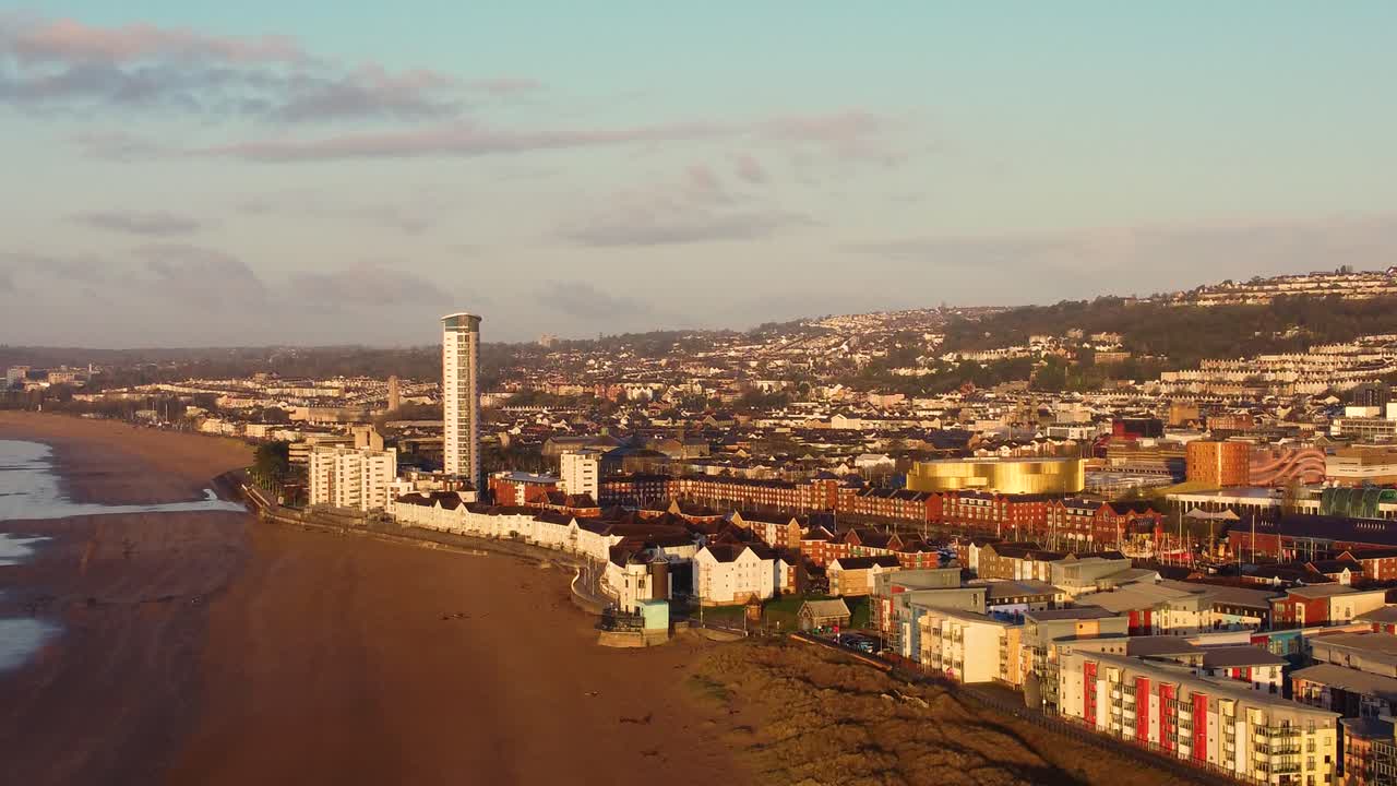 Ripresa aerea laterale del paesaggio urbano di Swansea con l'edificio più alto del Galles, appartamenti sul lungomare e l'Arena del centro città. Paesaggio urbano all'alba.