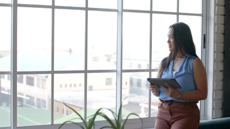 Una-Mujer-De-Negocios-Asiática-Está-Junto-A-La-Ventana-Sosteniendo-Una-Tableta,-Con-Espacio-Para-Copiar