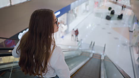 rear view of a young adult with long, flowing hair descending an escalator in a busy mall, she gracefully places her hand on the rail while observing the surroundings