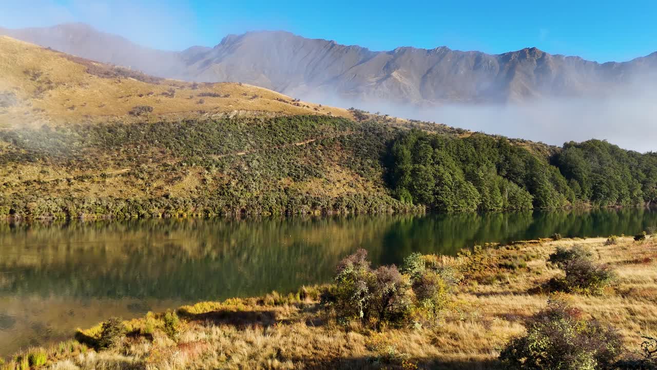 Aerial view of a serene mountain lake surrounded by mist and lush greenery during sunrise, captured by a drone