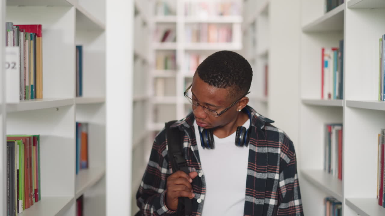 Black man looks around in public library. African American visitor with glasses and headphones chooses books in store. Education materials storage