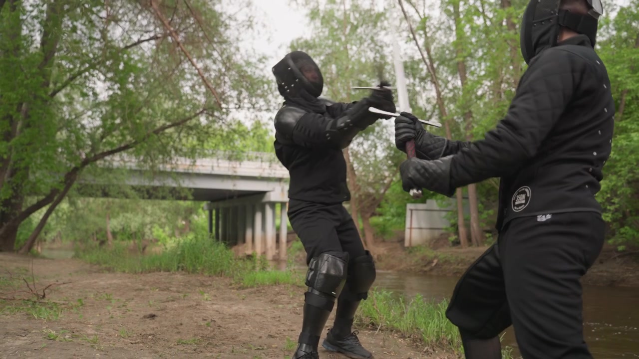 Close up of two people in black protective gear engaged in sword fight outdoors. One person holds weapon while another approaches with trees and overpass visible in background. Ground