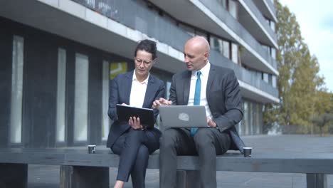 Business colleagues with laptop and tablet talking and working on project together while sitting at office building outside