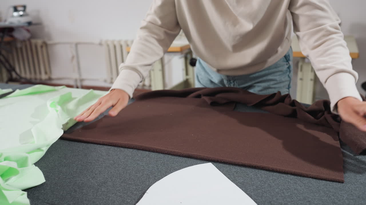 Female stylist smoothens brown material on work table preparing surface to place white cut out design shape during garment production process inside bright sewing studio surrounded by fabrics