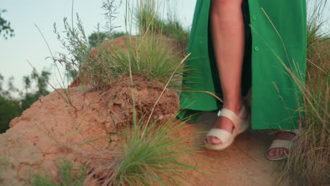lower angle view of woman in green gown stepping carefully down sandy hill trail surrounded by tall wild grass, earth texture visible beneath feet