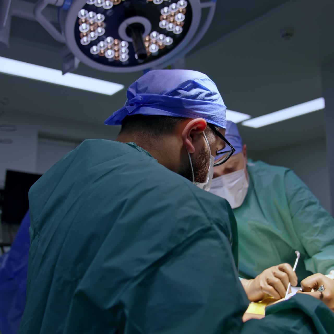 One surgeon widens the surgical section with metal tools. Main surgeon uses tools to operate the area. Anesthesiologist approaches the operational table addressing the doctor