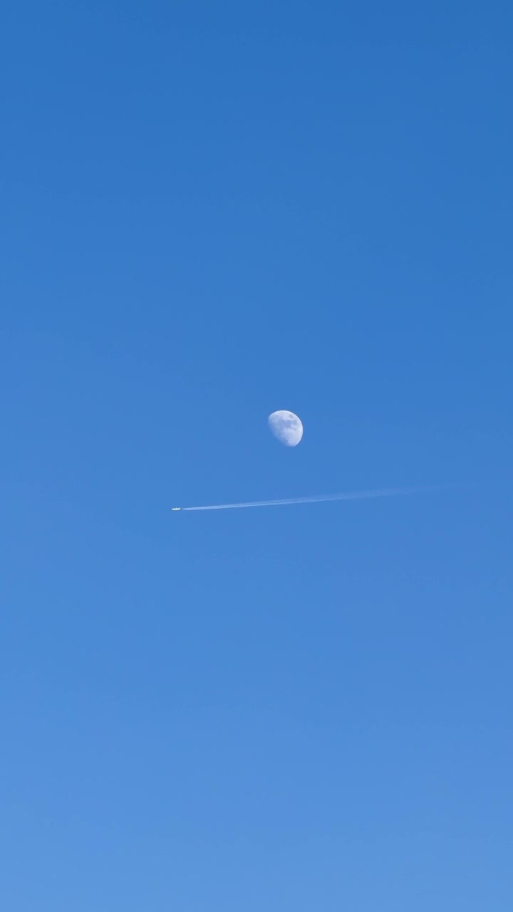 Un aereo lascia una scia di condensazione passando sotto la luna visibile in un cielo diurno limpido e blu.