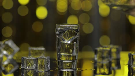 Barmann-Gießt-Gefrorenen-Wodka-Aus-Der-Flasche-In-Ein-Schnapsglas.-Eiswürfel-Vor-Goldglänzendem-Partyhintergrund