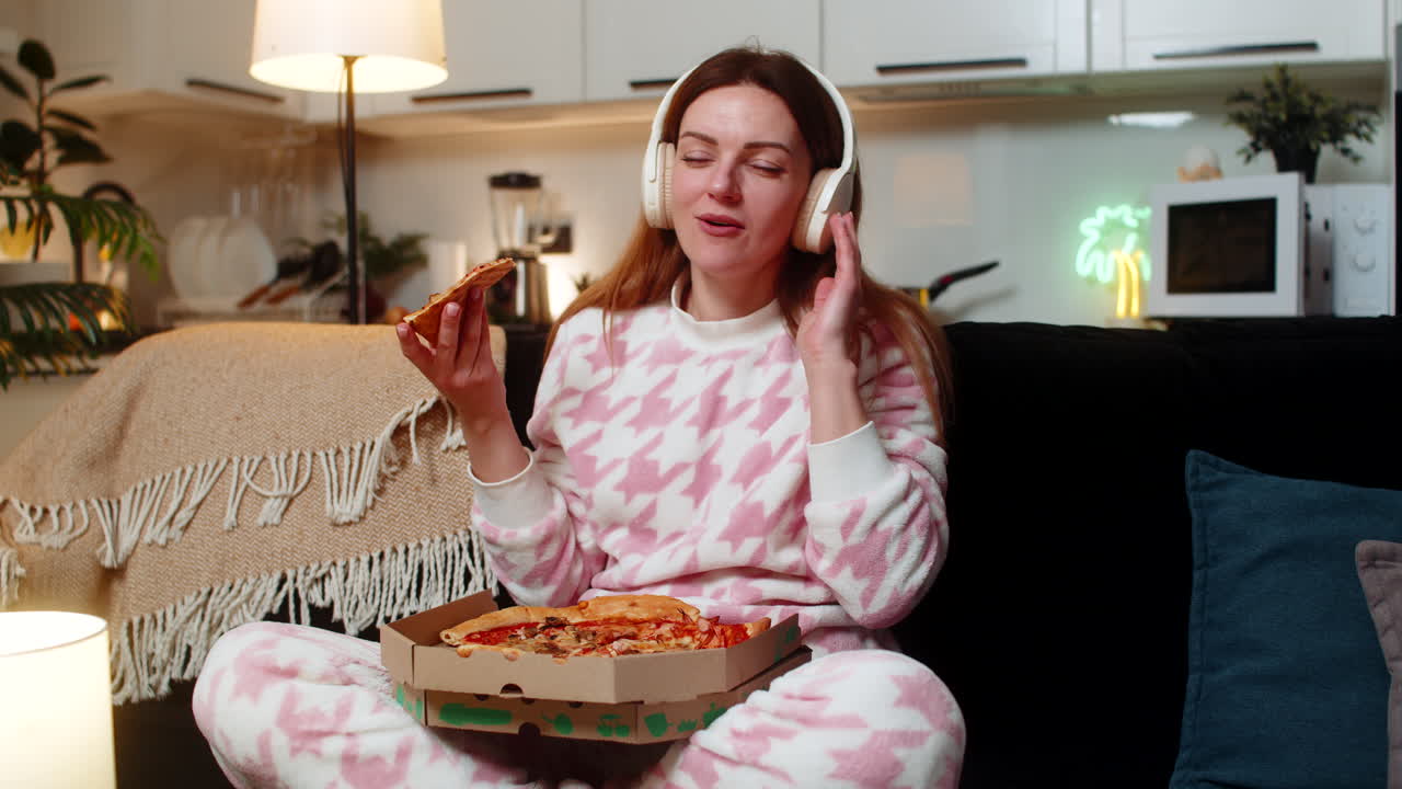 Adult woman dances with headphones at night enjoys music eats pizza smiles freedom happy cozy moment