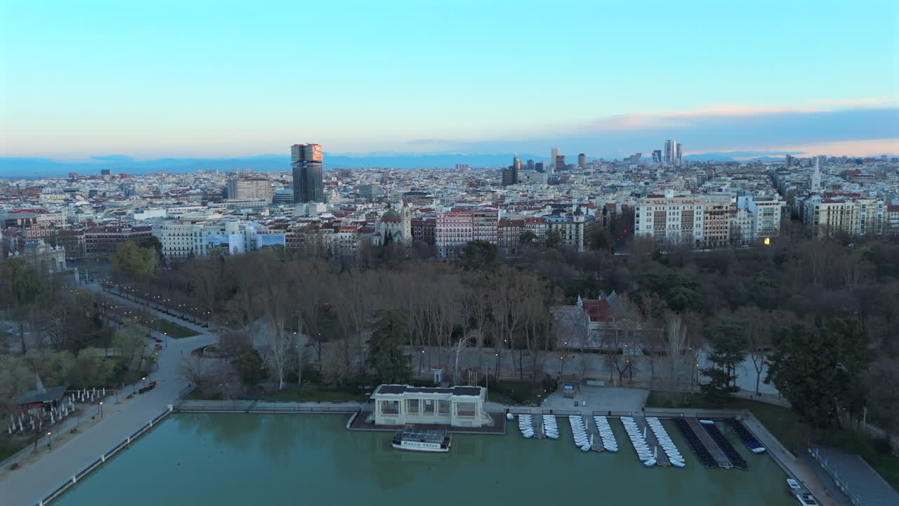 Drone footage flying backwards over a vibrant urban scene in Madrid. The clip reveals Retiro lake and lush park surroundings with early morning light and detailed architecture
