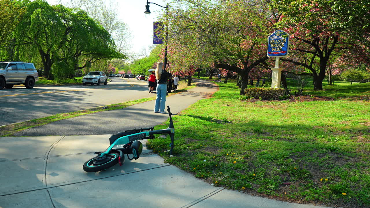 Nueva York, EE. UU., 2 de abril de 2026: Entrada del Parque Branch Brook con patinete eléctrico caído y árboles. Vista del letrero de la entrada del parque con un patinete eléctrico abandonado en la acera y cerezos en flor