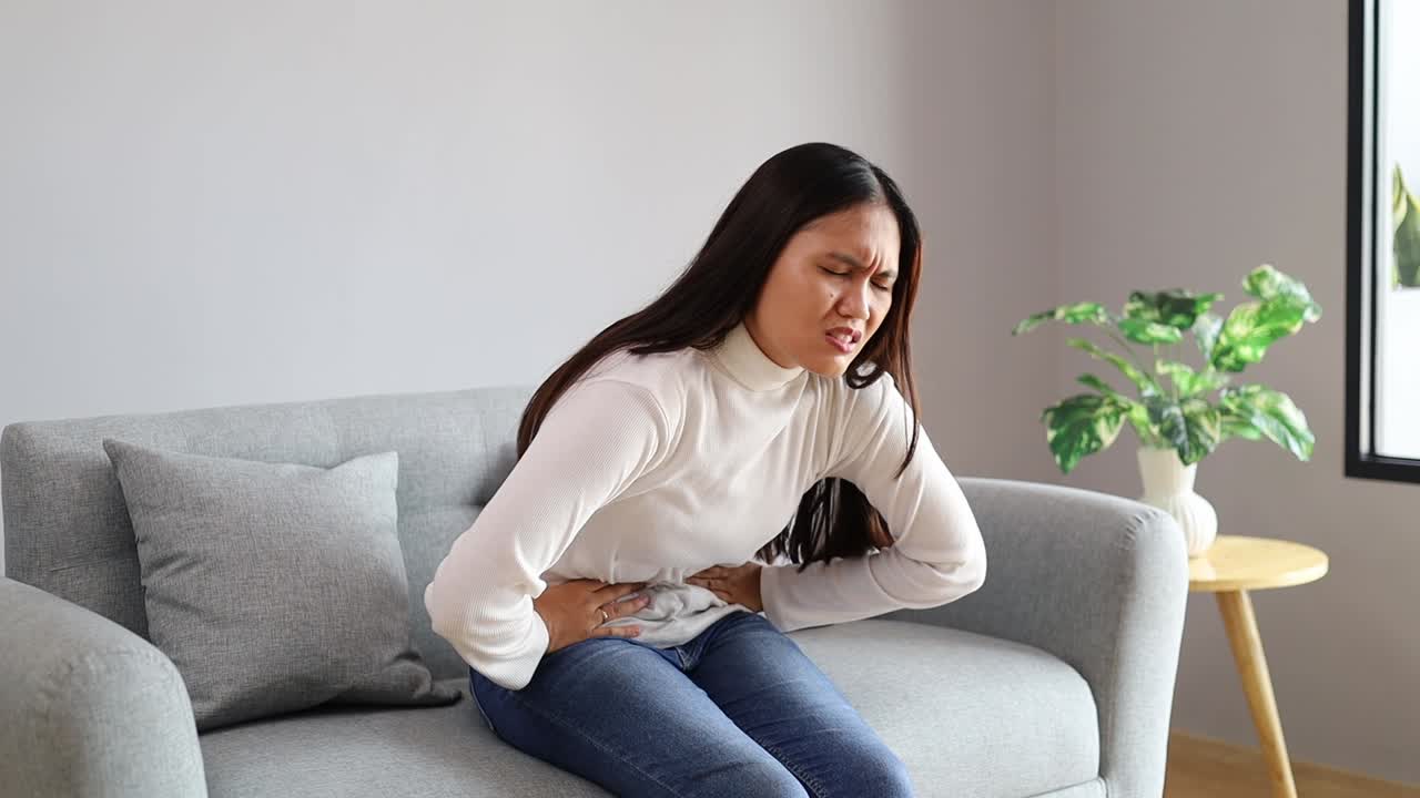 Young woman sitting on couch is experiencing intense abdominal pain. She is holding her belly and grimacing in discomfort