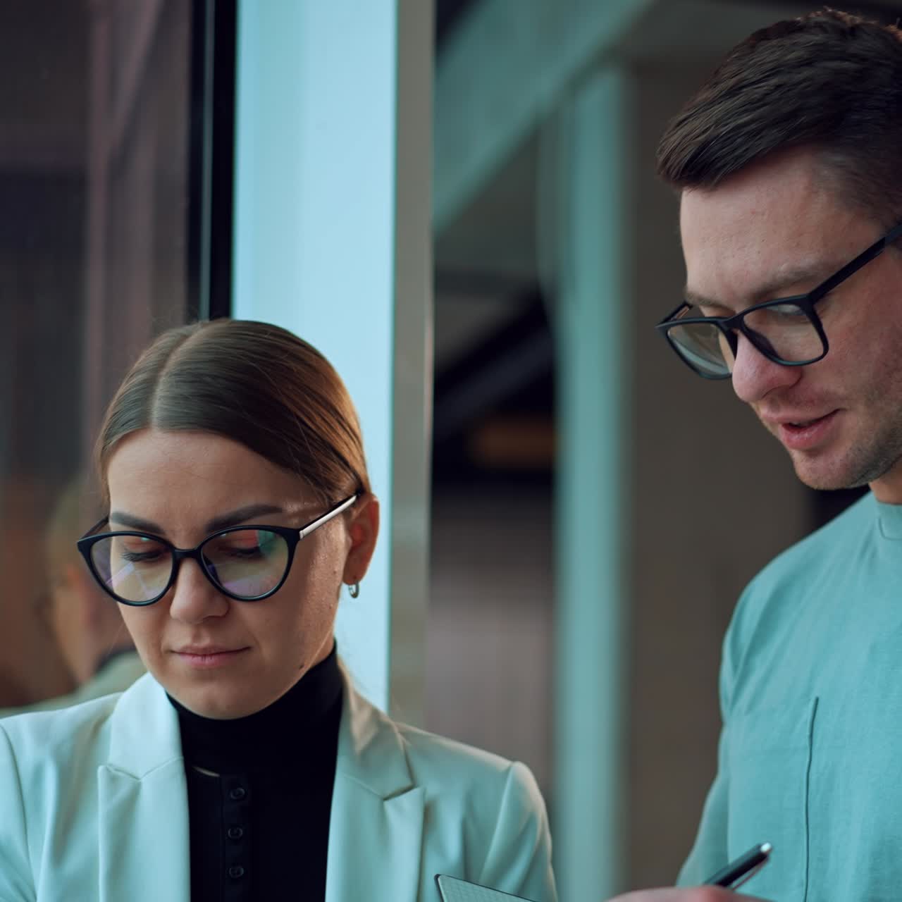 Young people working in the office discussing job issues standing at the window. Colleagues wearing glasses look at laptop talking about business