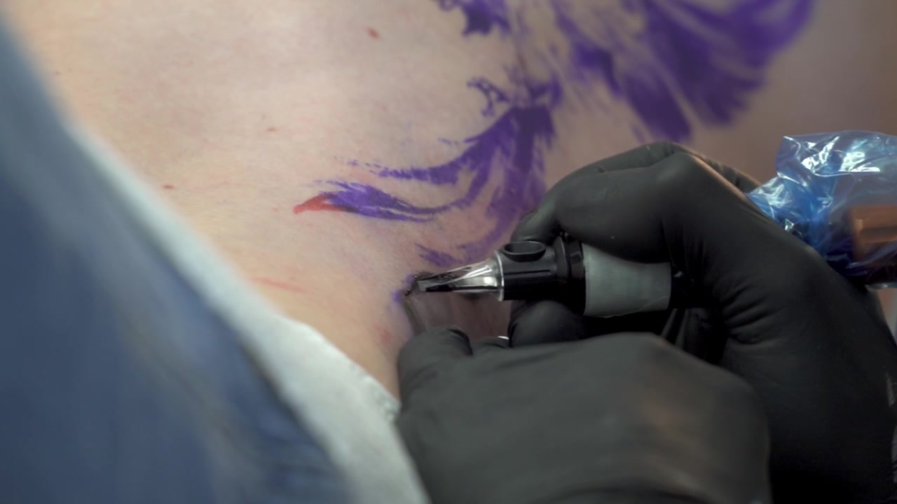 Un tatuatore con guanti neri inchiostra con precisione un disegno vivace usando una macchinetta per tatuaggi. Un primo piano evidenzia il dettaglio dell'ago e la maestria artistica.