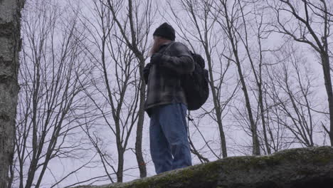 un hombre con una camisa de franela que lleva una mochila negra se encuentra en el ápice de una roca y domina el bosque con árboles estériles en el fondo en un día gris de invierno