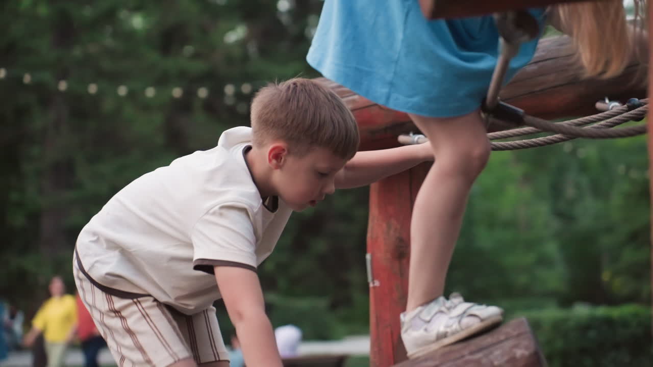 back view of boy climbing wooden playground structure while friend holds chain swing capturing summer adventure and teamwork in leafy green park setting at dusk with playful energy and exploration