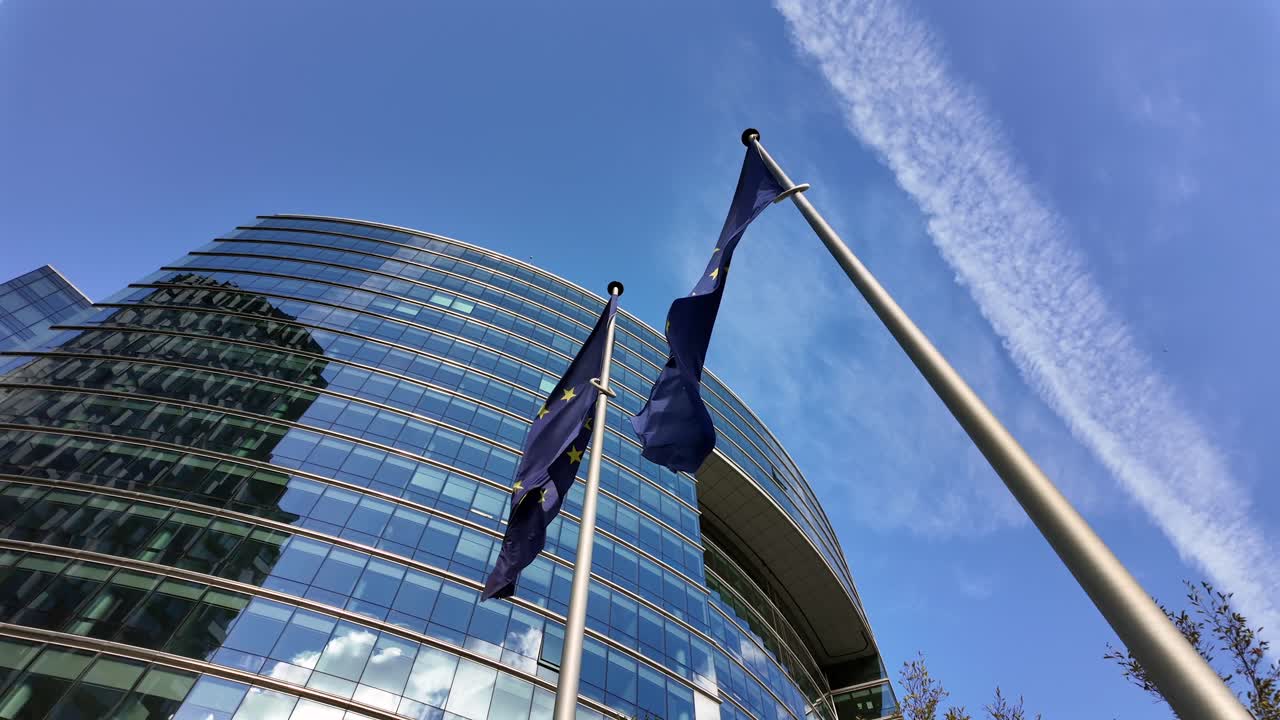 Bandiere dell'Unione Europea che sventolano di fronte al Lex Building a Bruxelles, con una scia di condensa di un aereo che attraversa il cielo azzurro.