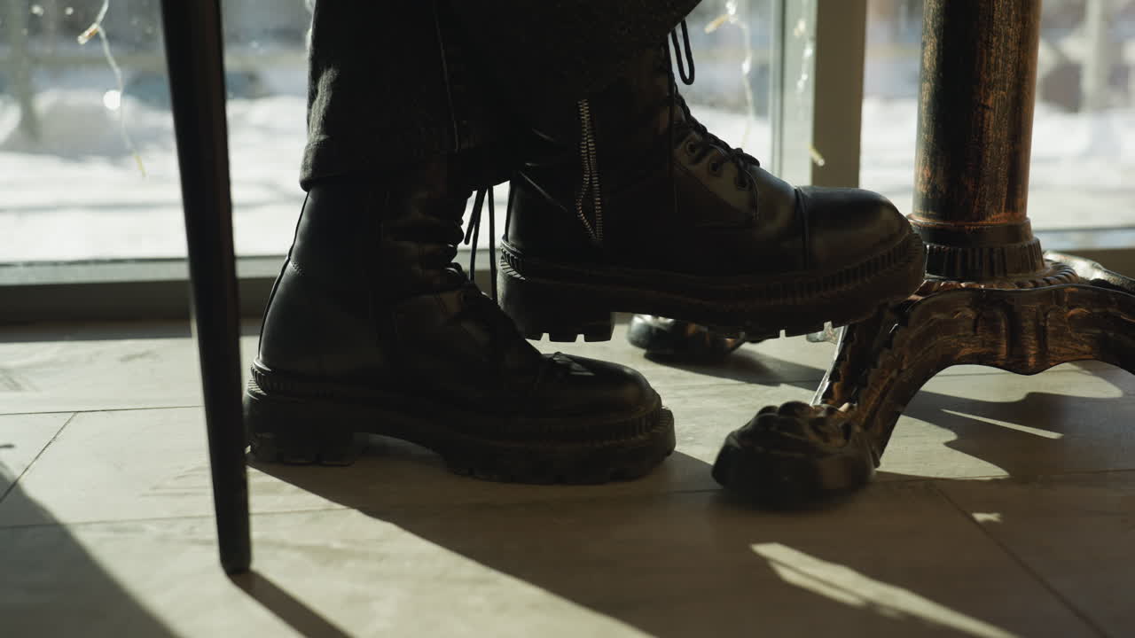 Leg view of lady seated at cafe table wearing black lace-up boots beside ornate metal table leg, with soft sunlight casting shadows on tiled floor and snowy street visible through glass panel