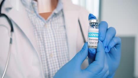 close up shot of healthcare worker specialist in protective uniform holding in hands in gloves ampoule with coronavirus vaccine, covid 19 vaccination concept, virus cure medicament, healthcare