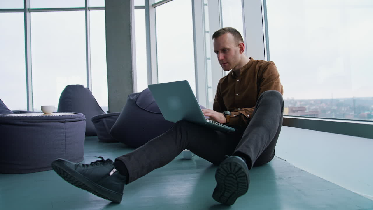 Young man sitting on floor near the window with a laptop. Happy man winning something while using a wireless computer.