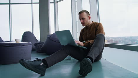 young man sitting on floor near the window with a laptop. happy man winning something while using a wireless computer.