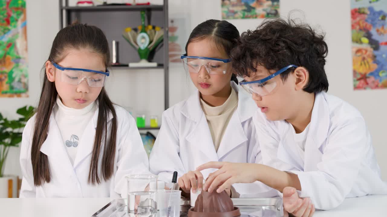 白衣と安全ゴーグルを身につけた3人のアジア系の子どもたちが、授業で化学実験を行っています。