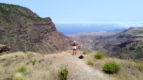 Tourist-girl-at-mountain-top-oustretched-with-open-arms-drone-aerial-view-of-valley-and-scape-in-Gran-Canaria