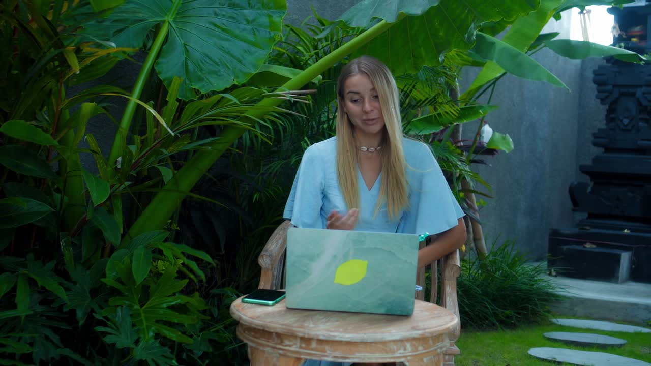 una chica en un vestido azul remotamente en línea trabajando detrás de una computadora portátil y mirando a la pantalla el patio trasero con plantas verdes