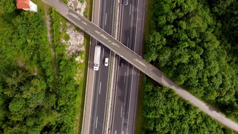 Top-down-drone-shot-of-lightly-wet-European-highway-with-overpass-and-power-lines-going-across-it-and-trees-surrounding-the-national-road