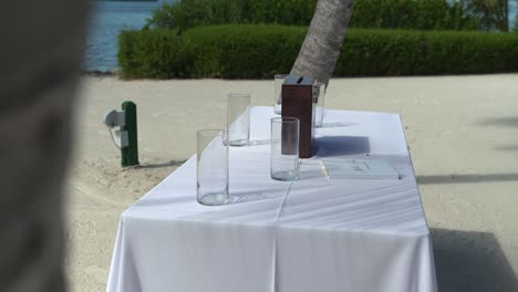 Toma-Panorámica-Desde-Un-árbol-Hasta-Una-Mesa-Con-Letrero-Lugar-De-Celebración-De-Bodas-Junto-A-La-Playa-En-Un-Día-Soleado-Con-Paz-Y-Tranquilidad-Asientos-Al-Aire-Libre-Playa-De-Arena-Asientos-Con-Mesas