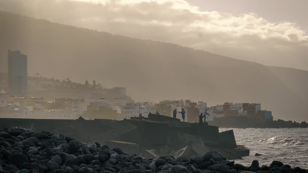 Men standing over the rocks at the shore just along the ocean, under a cloudy sky. Slow motion.