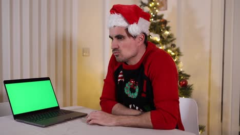Festlicher-Weihnachtsmann-Mit-Einem-Weihnachtspullover,-Der-Einen-Green-screen-laptop-Mit-Nachdenklichem-Ausdruck-Beobachtet,-Der-Vor-Einem-Geschmückten-Weihnachtsbaum-Mit-Den-Fingern-Auf-Den-Tisch-Klopft