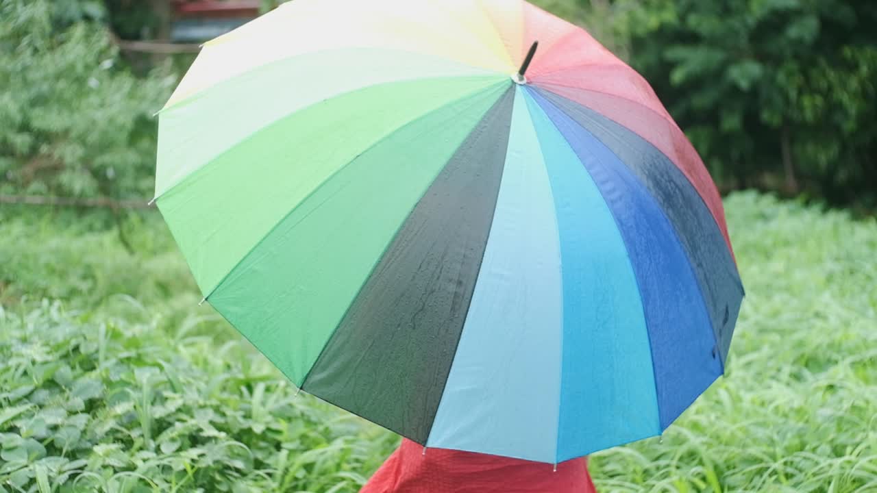 mujer caucásica de piel pálida vestida de rojo caminando en el jardín bajo la lluvia con un paraguas arcoiris en cámara lenta