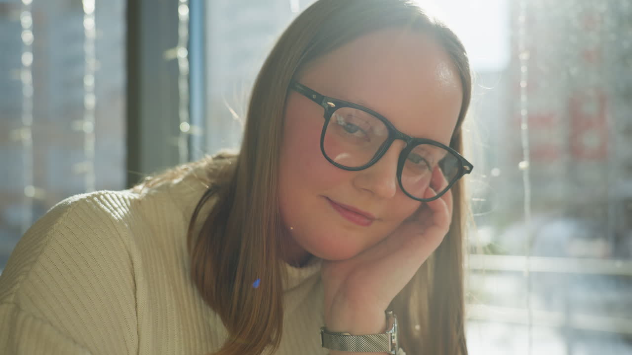 Calm student with glasses rests head on hand while gazing thoughtfully at camera in cozy indoor space, sunlight streams through window decorated with string lights, creating soft background glow