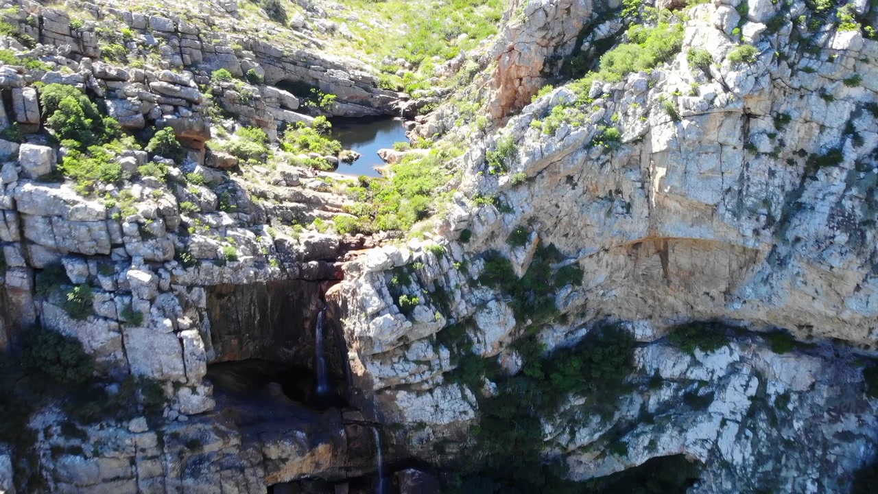 Steile Felsformationen mit dunklen Höhlenöffnungen, einem kleinen Wasserfall, der von einem zurückgesetzten Felsvorsprung herabstürzt, und Flecken grüner Vegetation unter klarem natürlichem Licht.