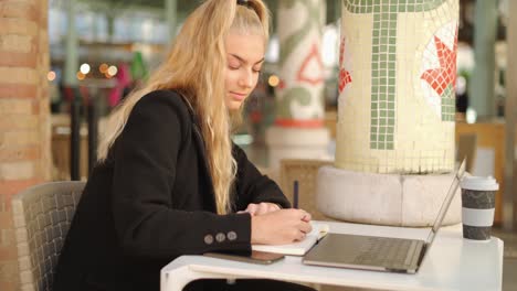 freelancer woman writing notes while working with laptop in cafe