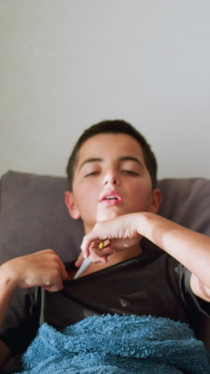 Sick boy sitting on couch collects thermometer from sister to check his temperature, placing it under his armpit while covered with a cozy blanket, soft natural light filters through the window