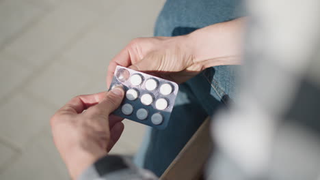 Close up of person in jeans carefully pressing tablet from blister medicine pack while seated on wooden bench in outdoor daylight setting with pavement pattern and subtle clothing