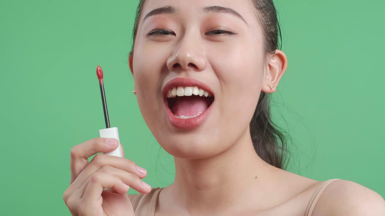 미소 짓는 아시아 여성이 립스틱을 들고 녹색 화면 배경에 서서 카메라에 키스를 보내고 있습니다.