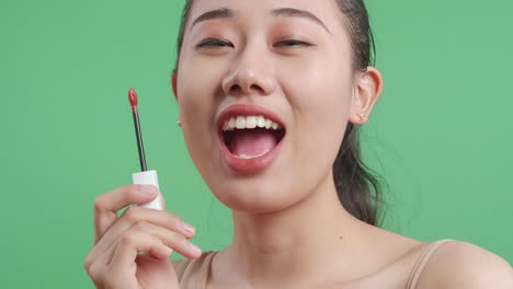 un primer plano de una mujer asiática sonriente sosteniendo lápiz labial y enviando un beso a la cámara mientras está de pie en el fondo de la pantalla verde en el estudio