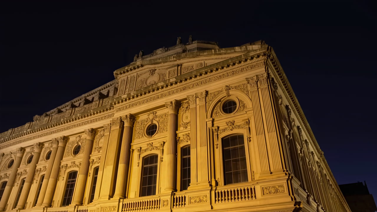 Neoclassical Facade Lit At Night Showcasing Ornate Columns, Pilasters, Cornices, And Balconies Bathed In Warm