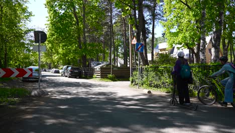 children driving on a small road in nõmme with an electric scooter and a bicycle during summer time and turning to a different street. one way ending traffic sign, trees and fences visible in sunshine
